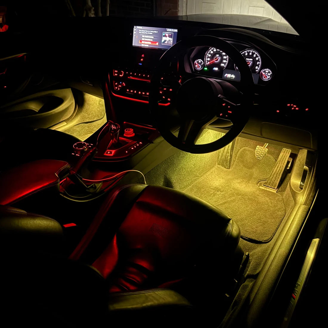 A car interior illuminated by warm yellow under-seat and door light strips, showing black leather seats, a steering wheel, dashboard, and carpeted floor with red ambient accent lighting.
