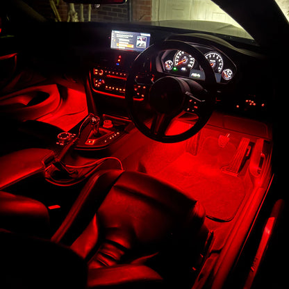 A car interior illuminated by bright red ambient lighting, showing black leather seats, a black steering wheel, dashboard instruments, and red light reflecting across the floor and console.