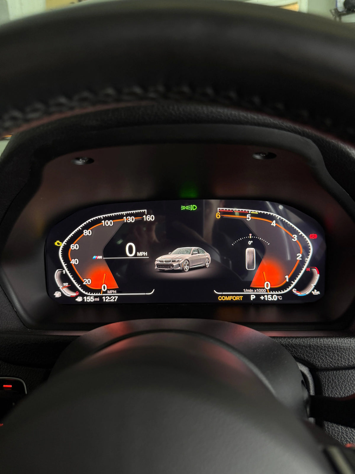A close-up view of a BMW's digital instrument cluster display retrofit, showing a silver sedan model graphic in the center, with black steering wheel and dashboard surrounding the screen.