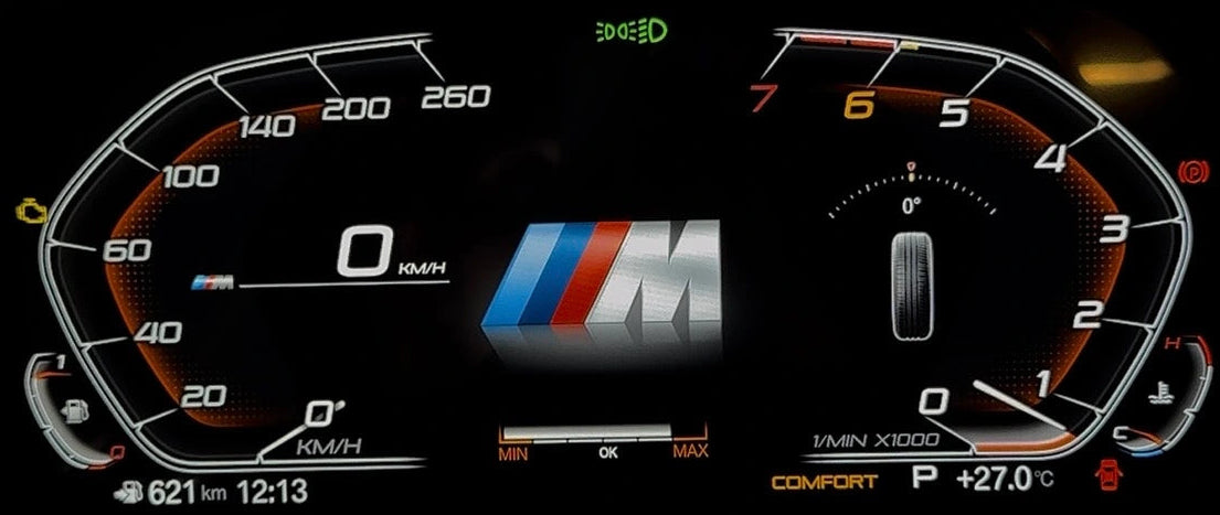 A BMW E90 digital instrument cluster display on a car dashboard, showing illuminated speedometer, tachometer, and center M logo with orange and white markings on a black screen.