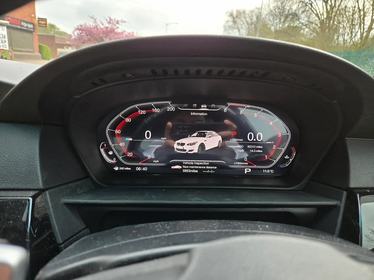 A close-up view of a BMW's digital instrument cluster display, showing a white E60 car graphic and speedometer gauges, installed inside a black dashboard.