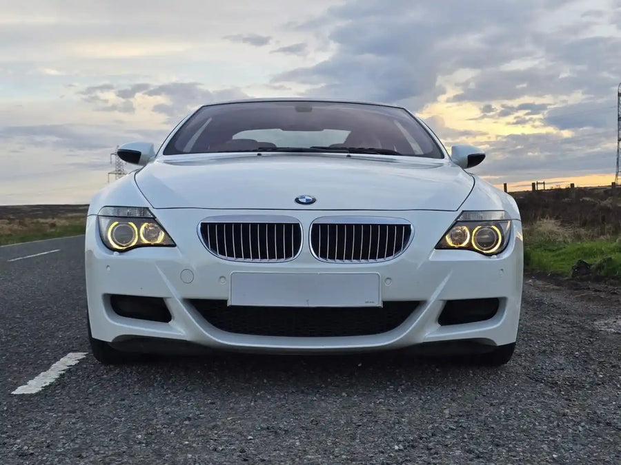 A white BMW M6 coupe is parked on a paved road at dusk, facing the camera. The car features its signature kidney grille and illuminated halo headlights.