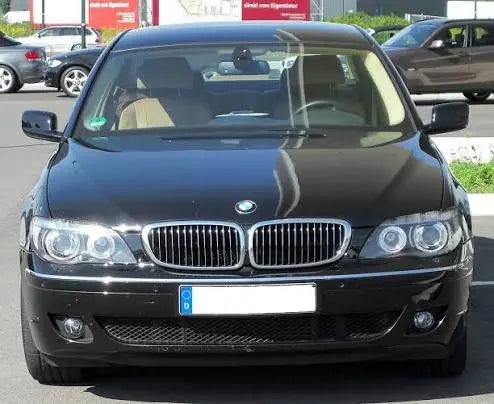 A front-facing black BMW e65 7 series sedan parked outdoors in a parking lot. The car shows the iconic BMW kidney grille, clear headlights, and a blank white license plate area.