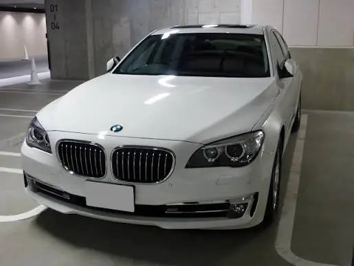 A white BMW F01 7 series sedan parked in an indoor parking structure. The front of the car is visible, showing the iconic BMW kidney grille, headlights, and a blank license plate area.