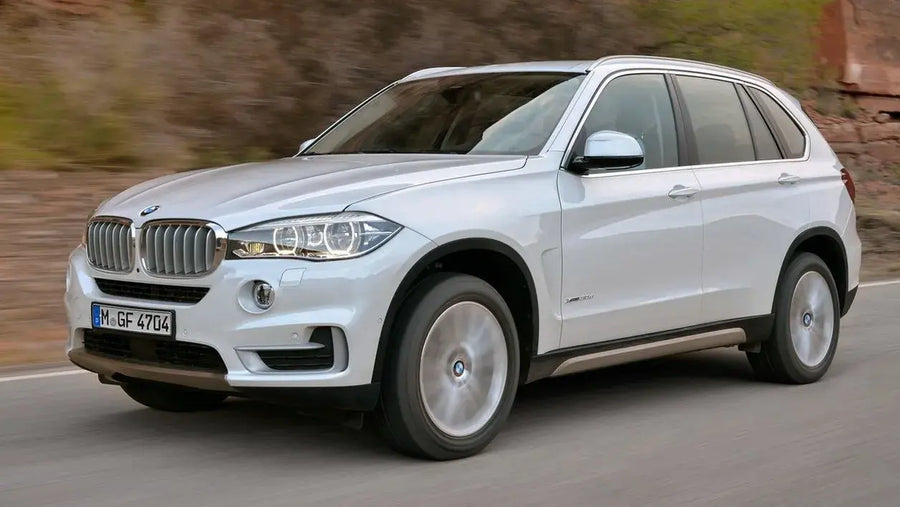 A silver BMW X5 F15 SUV captured in motion on a paved road, showing its front grille, headlights, side profile, and wheels against a blurred outdoor background.