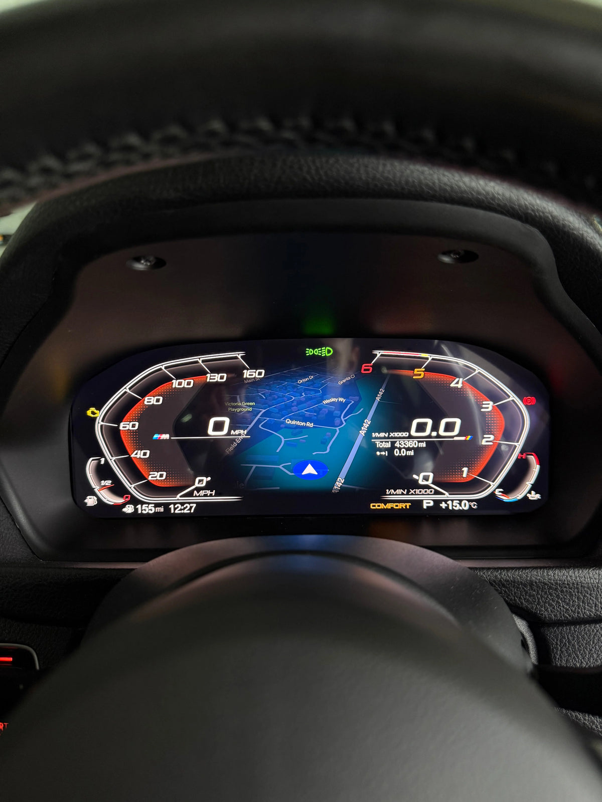 Close-up of a BMW's digital instrument cluster display showing a blue and red dashboard interface with carplay maps, framed by a dark leather steering wheel and dashboard.