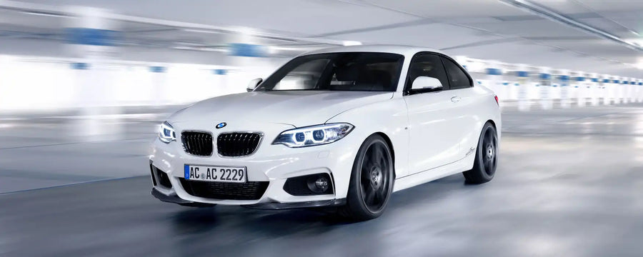A white BMW 2 Series coupe is shown driving in a blurred indoor parking structure. The car features the iconic BMW kidney grilles, blue LED headlights, and black multi-spoke wheels.