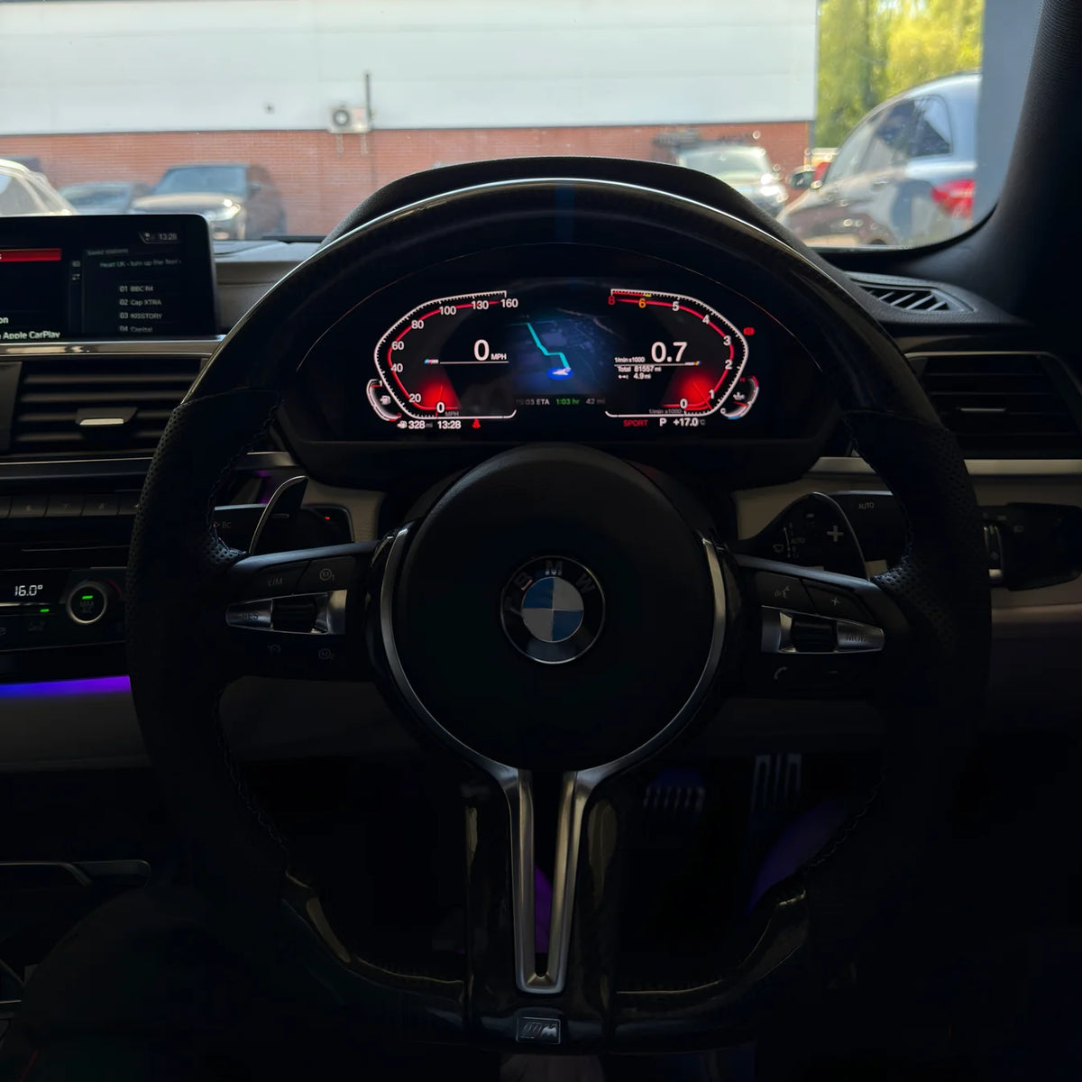A close-up view of a black BMW M-style steering wheel with a retrofitted digital instrument cluster behind it, photographed inside a car.