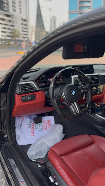 A BMW car interior with red leather seats and red trim, viewed from the open driver's side door. The steering wheel has the BMW logo, and the speedometer is retrofitted to digital.