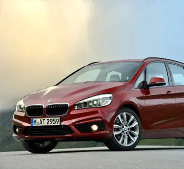 A red BMW 2 Series Active Tourer station wagon shown from a front three-quarter angle, parked outdoors on a paved surface with a soft-focus sky and landscape background.
