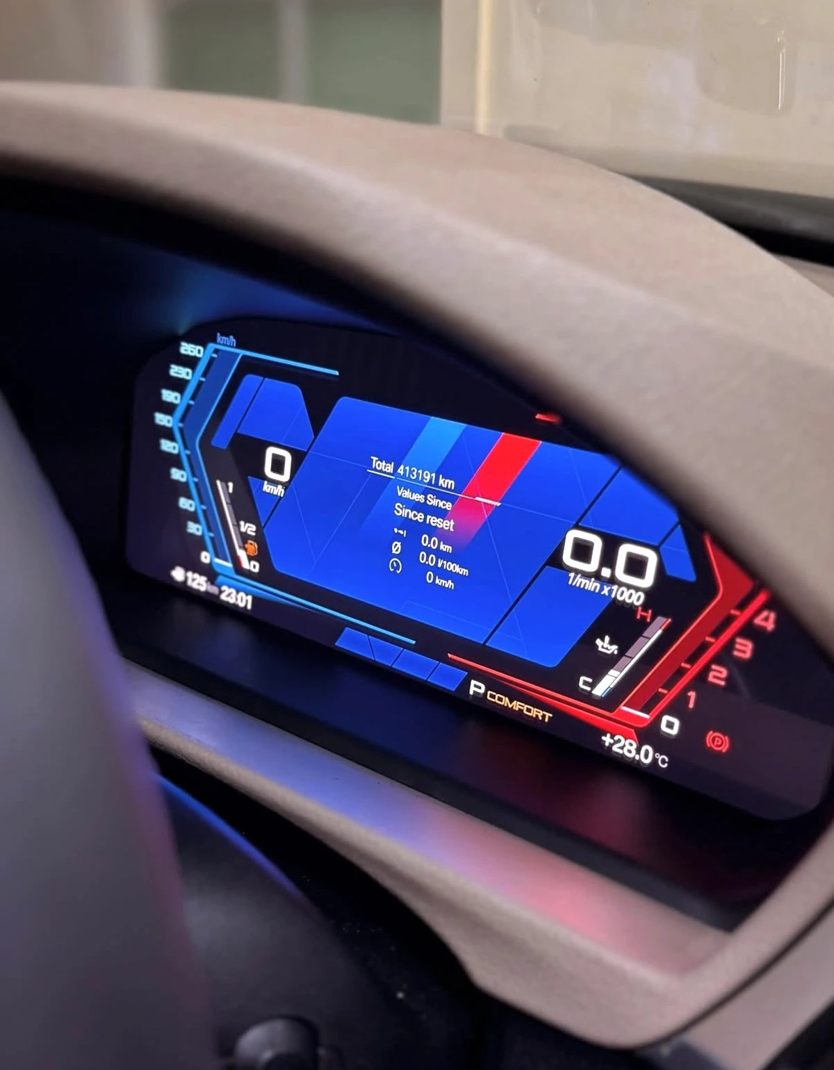 Close-up of a BMW digital instrument cluster display retrofit showing blue, red, and white interface graphics on a dark dashboard, framed by a beige steering wheel.