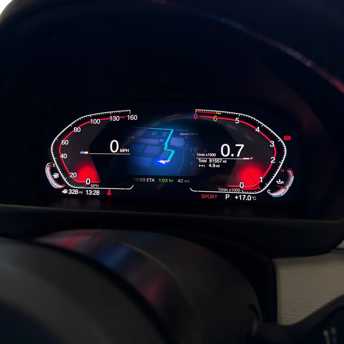 A close-up of a BMW's digital instrument cluster display showing a dark dashboard interface with red, white, and blue illuminated graphics and speedometer/tachometer markings and maps.
