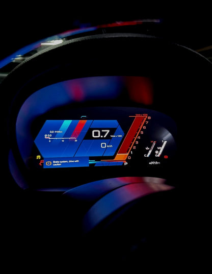 A close-up of a BMW's digital instrument cluster display upgrade illuminated in dark tones, showing blue, red, and orange dashboard graphics and vehicle metrics.