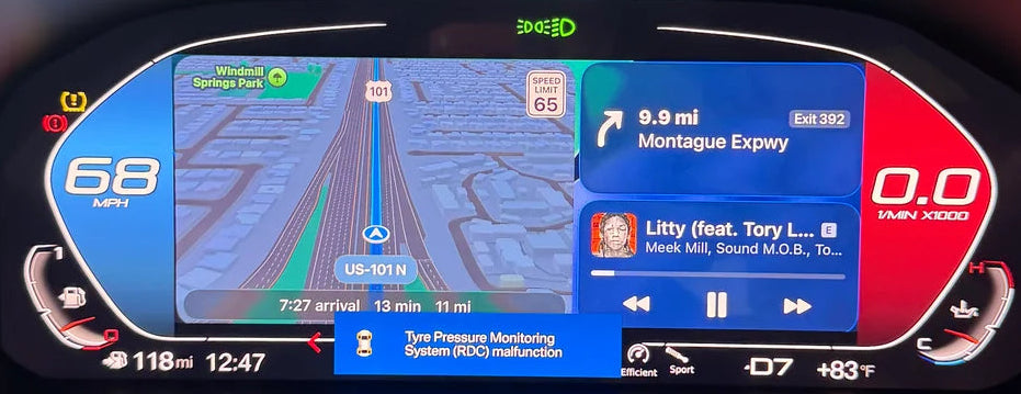 A close-up of a BMW's digital instrument cluster display showing navigation and media, with a blurred plush toy in the foreground.