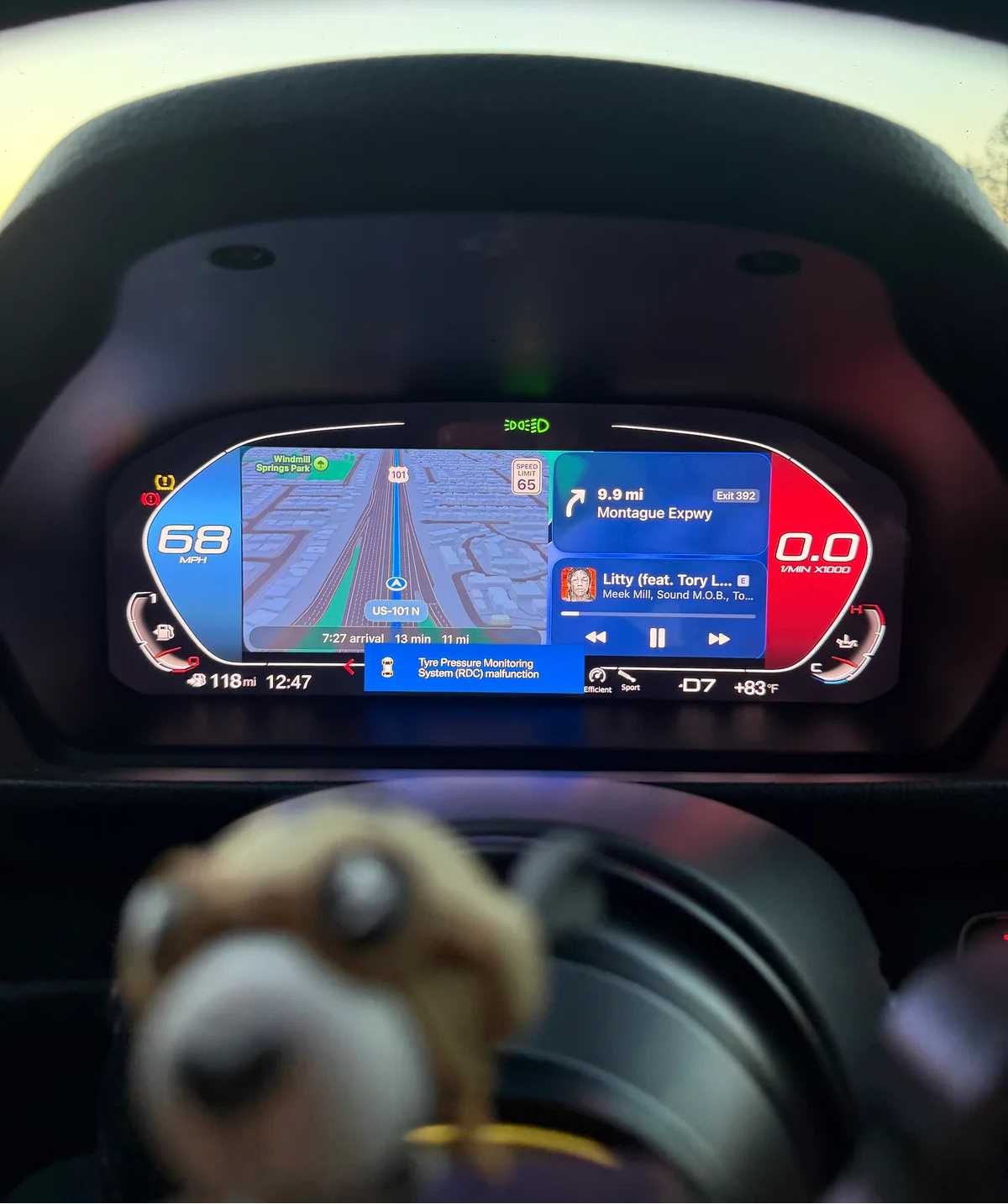 A close-up of a BMW's digital instrument cluster display showing navigation and media, with a blurred plush toy in the foreground.