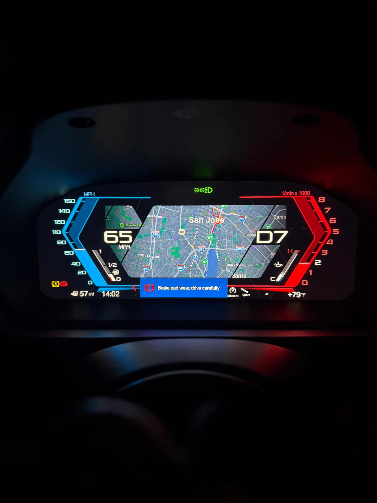 A close-up of a BMW digital instrument cluster display on a car dashboard, showing a navigation map, speed of 65 mph, and tachometer readings, illuminated with blue and red accents in a dark setting.
