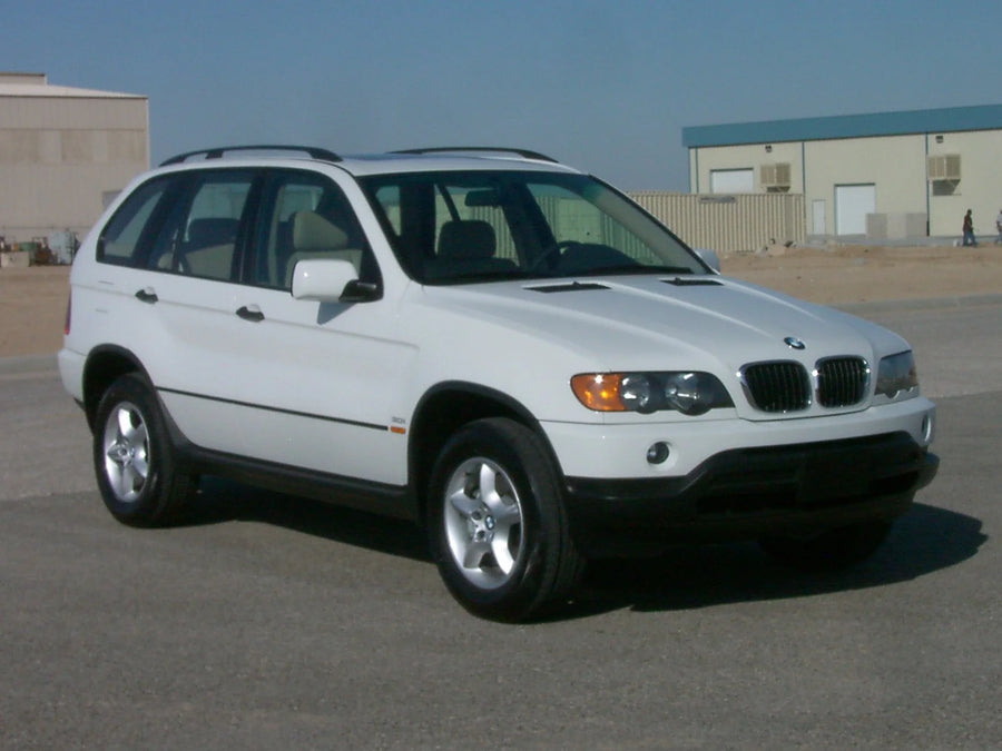 A white BMW X5 E53 SUV parked outdoors on a paved surface, with industrial buildings and a blue sky in the background.