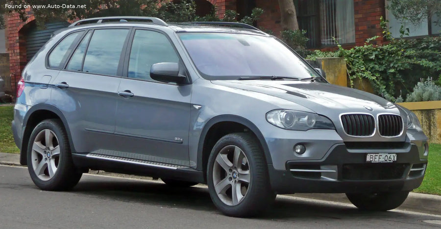 A silver BMW X5 E70 SUV parked on a street in front of a brick house with greenery.