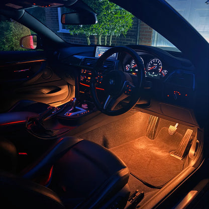 Underfloor car lighting illuminated in a dark vehicle interior, casting a warm orange glow over the carpet and pedals, with black seats and dashboard visible.