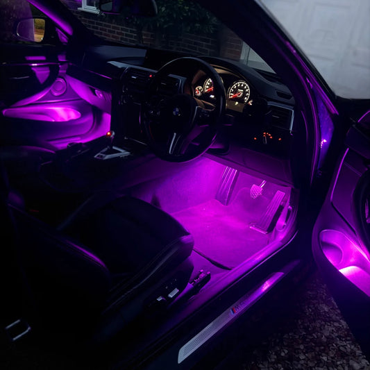 Purple LED car interior ambient light glowing in the cabin, illuminating the footwell, door panel, and steering wheel area of a black car.