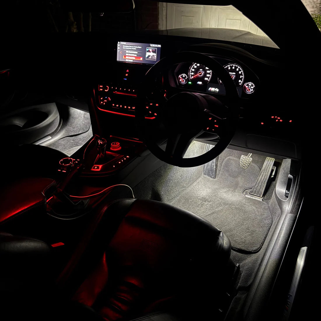 A car interior with illuminated red ambient lighting, showing the dashboard, steering wheel, center console, footwell, and dark leather seats.