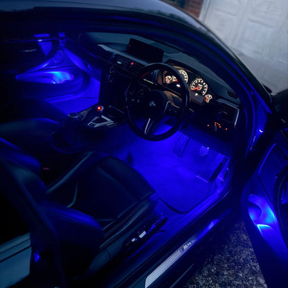 Blue LED under-seat car interior lighting illuminating a black leather cabin with an open driver's side door, showing the footwell and dashboard area.