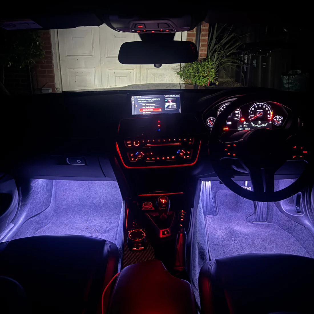 A car interior at night with illuminated purple footwells and red ambient lighting around the console, steering wheel, and dashboard. The windshield shows a residential garage and plants outside.