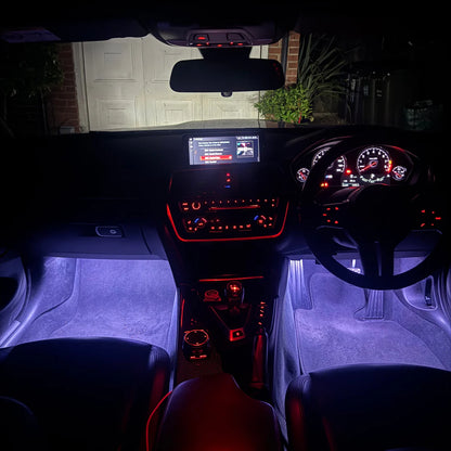 A car interior at night with illuminated purple footwells and red ambient lighting around the console, steering wheel, and dashboard. The windshield shows a residential garage and plants outside.
